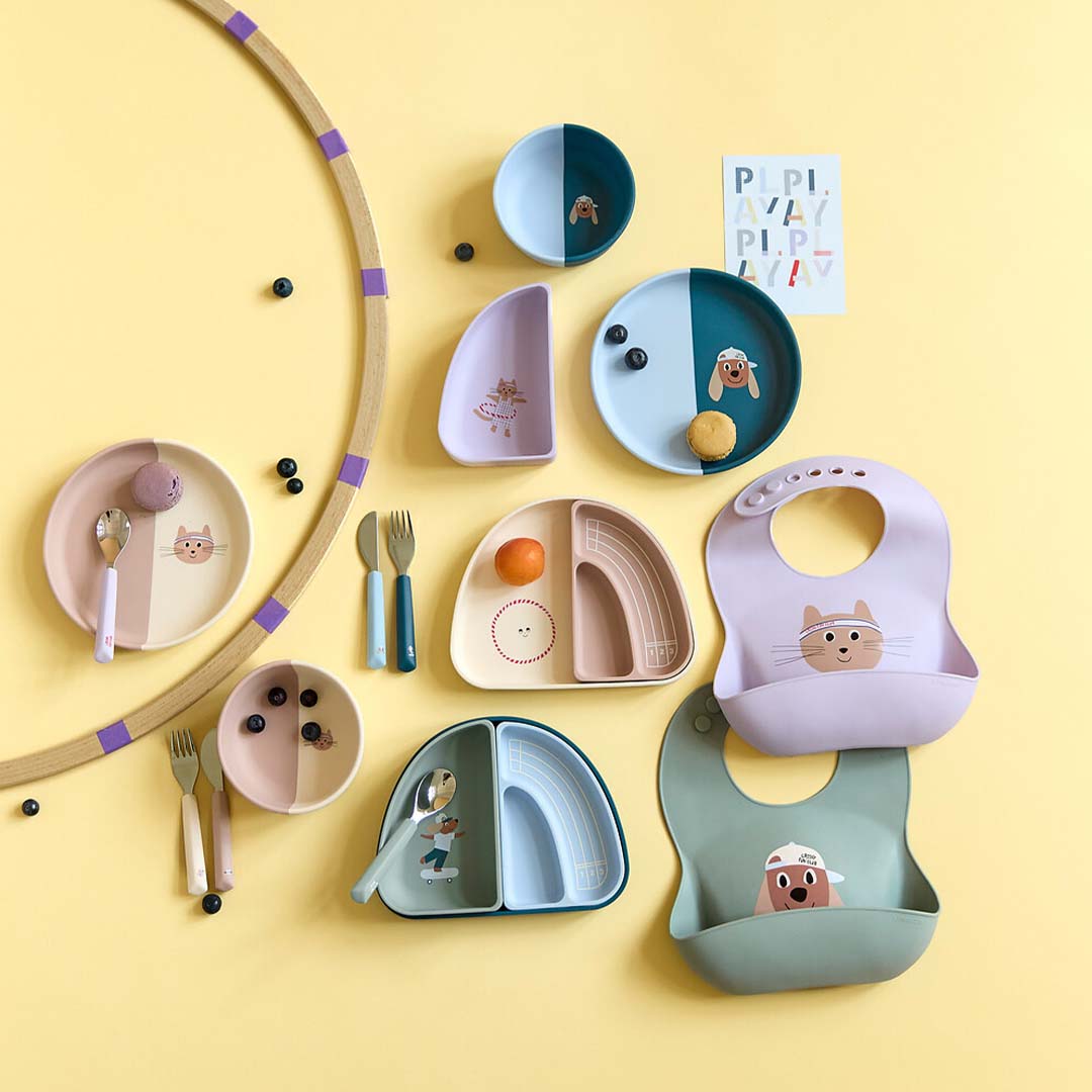 Een collectie Lässig baby voedingsbenodigdheden, waaronder paarse en groene siliconen slabbetjes met dierenfiguren, borden en bestek.