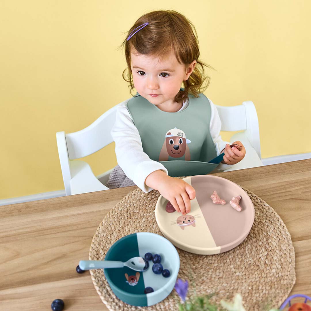 Een baby draagt het Lässig groene siliconen slabbetje met hondenthema aan tafel met een bord en kom.