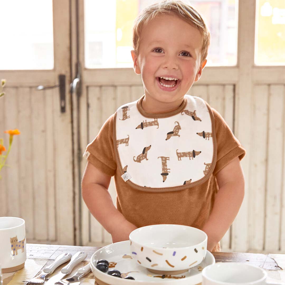 Lachende peuterjongen draagt een Lässig babyslabbetje met teckelpatroon aan een eettafel.