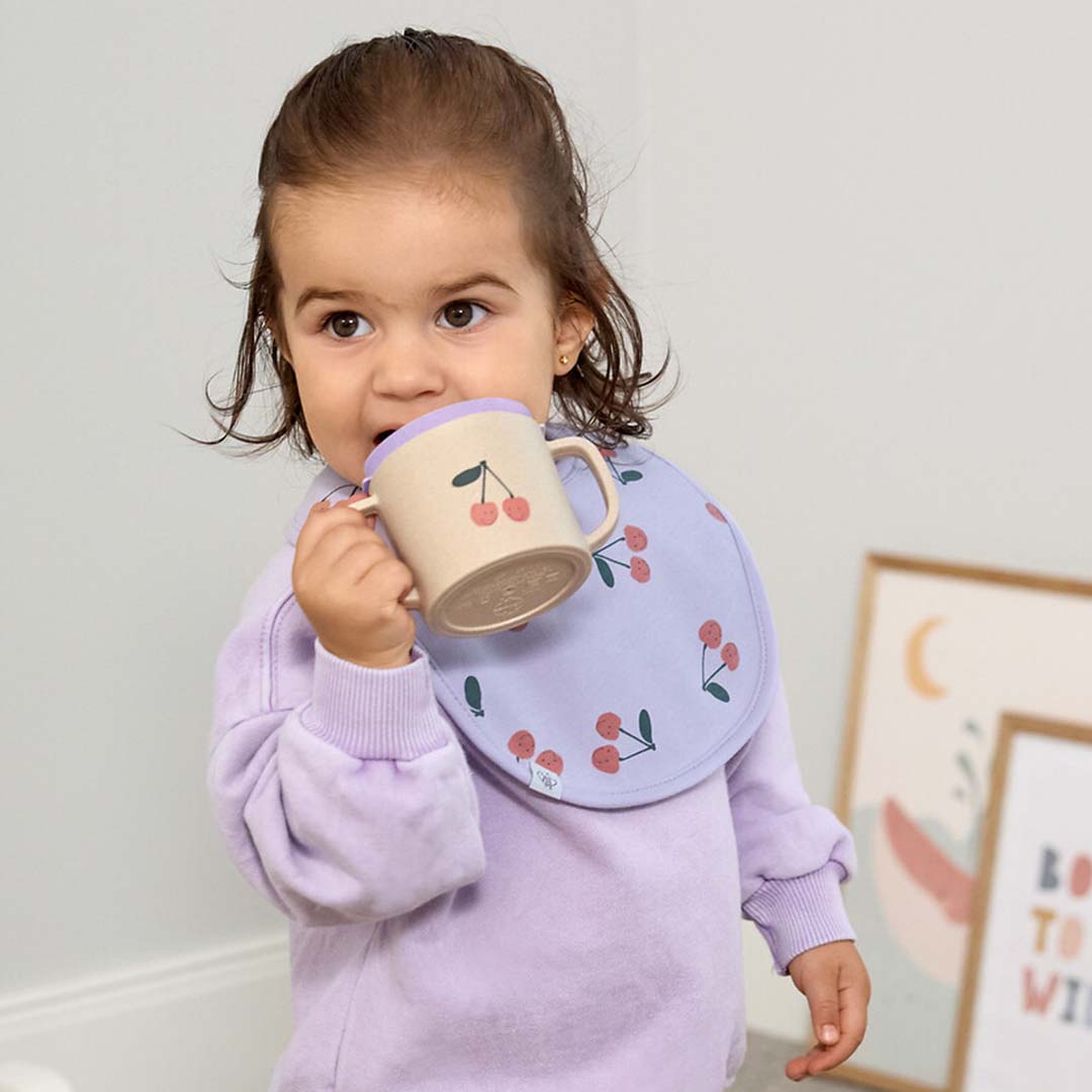 Een baby met een paarse slabber drinkt uit de Lässig tuitbeker met kersenpatroon.