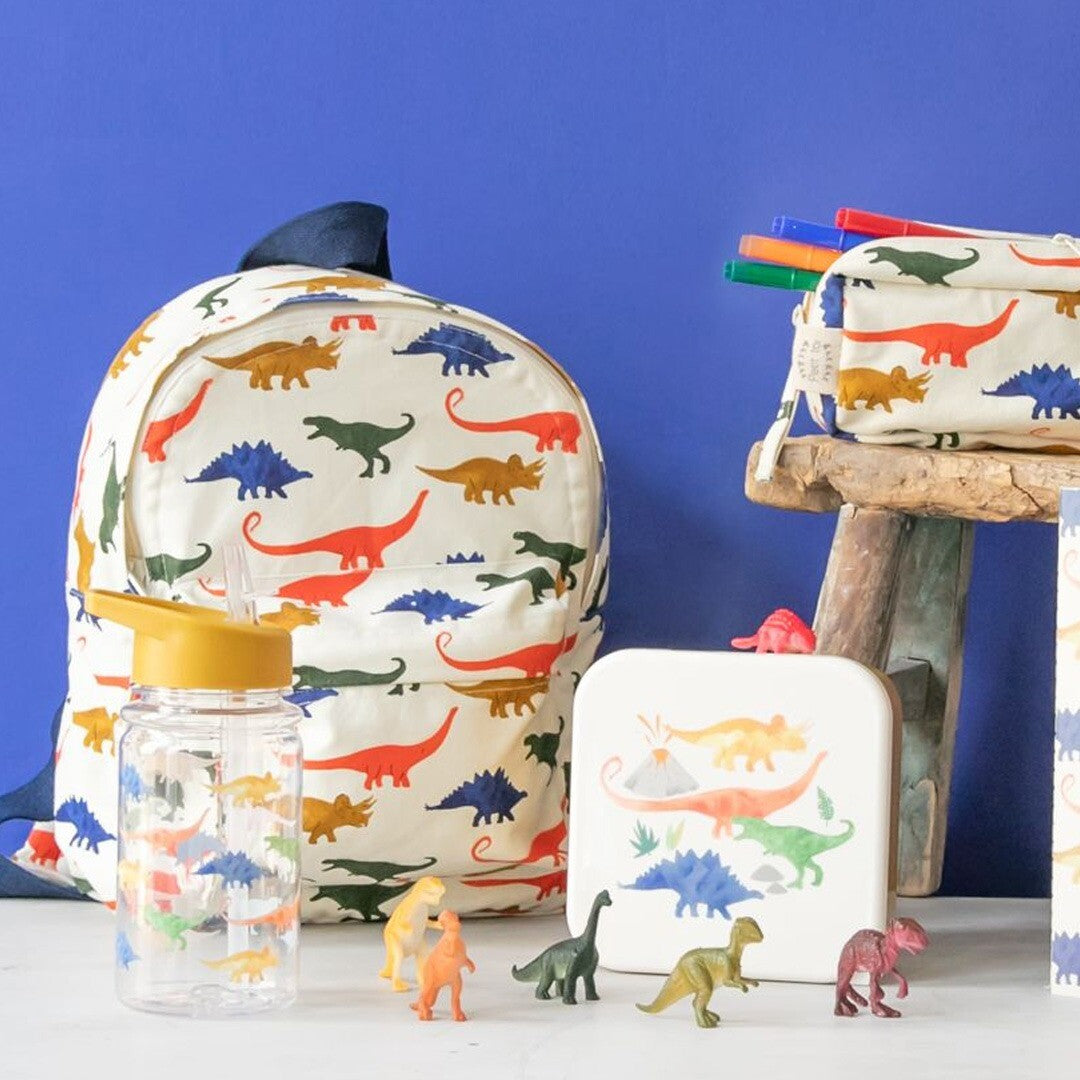 Le petit souk collectie van schoolspullen met dinosauruspatroon, inclusief een rugzak, drinkfles, etui en broodtrommel, omringd door speelgoeddinosaurussen.