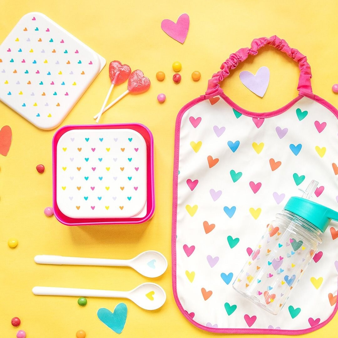 Een le petit souk lunchbox set, een slabbetje, een drinkfles en lepels met hartjespatroon op een gele achtergrond.