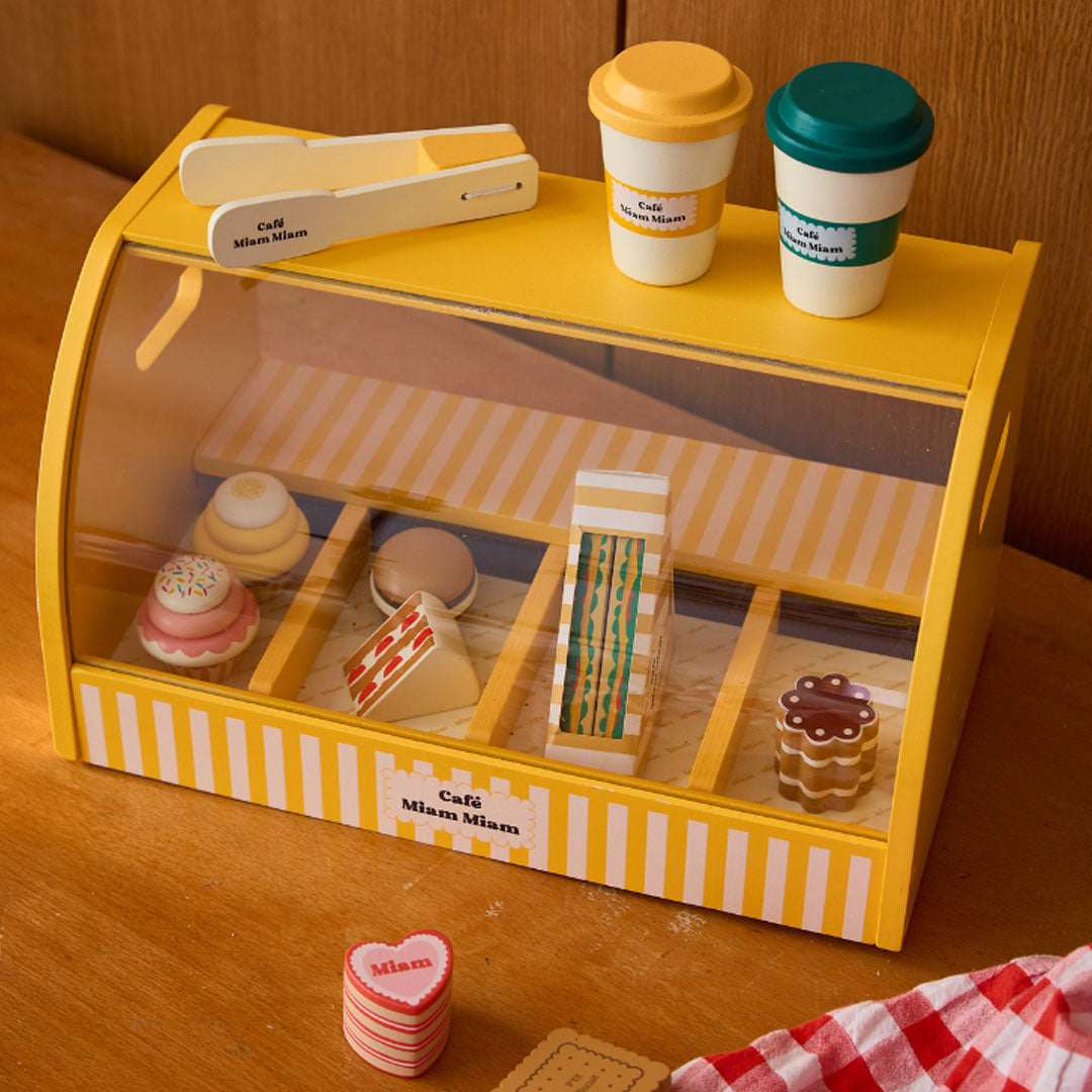 Le petit souk houten keukenspeelset met gele gestreepte toonbank, gevuld met speelgoedgebakjes en koffiekopjes op een houten tafel.