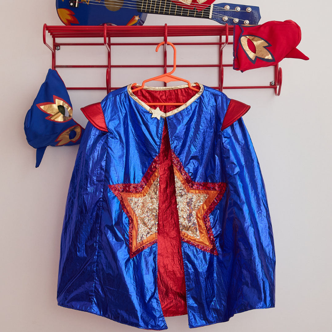 le petit souk superheldencape voor kinderen, blauw met rode binnenkant en een gouden en rode ster, hangend aan een rode kapstok met andere superheldenaccessoires.