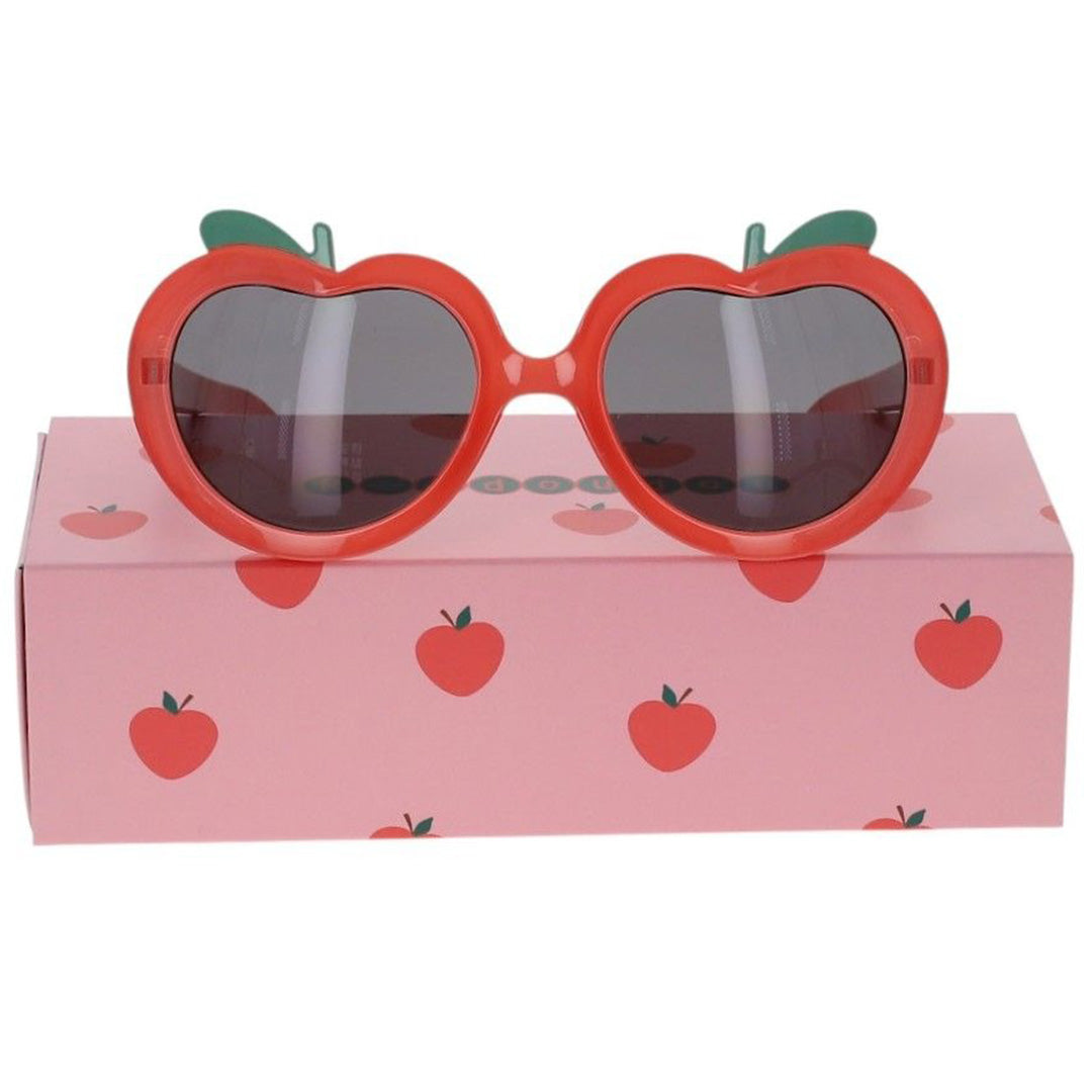 le petit souk kinderzonnebril met rood hartvormig montuur en groene steeltjes, liggend op de roze verpakking met appelprint.