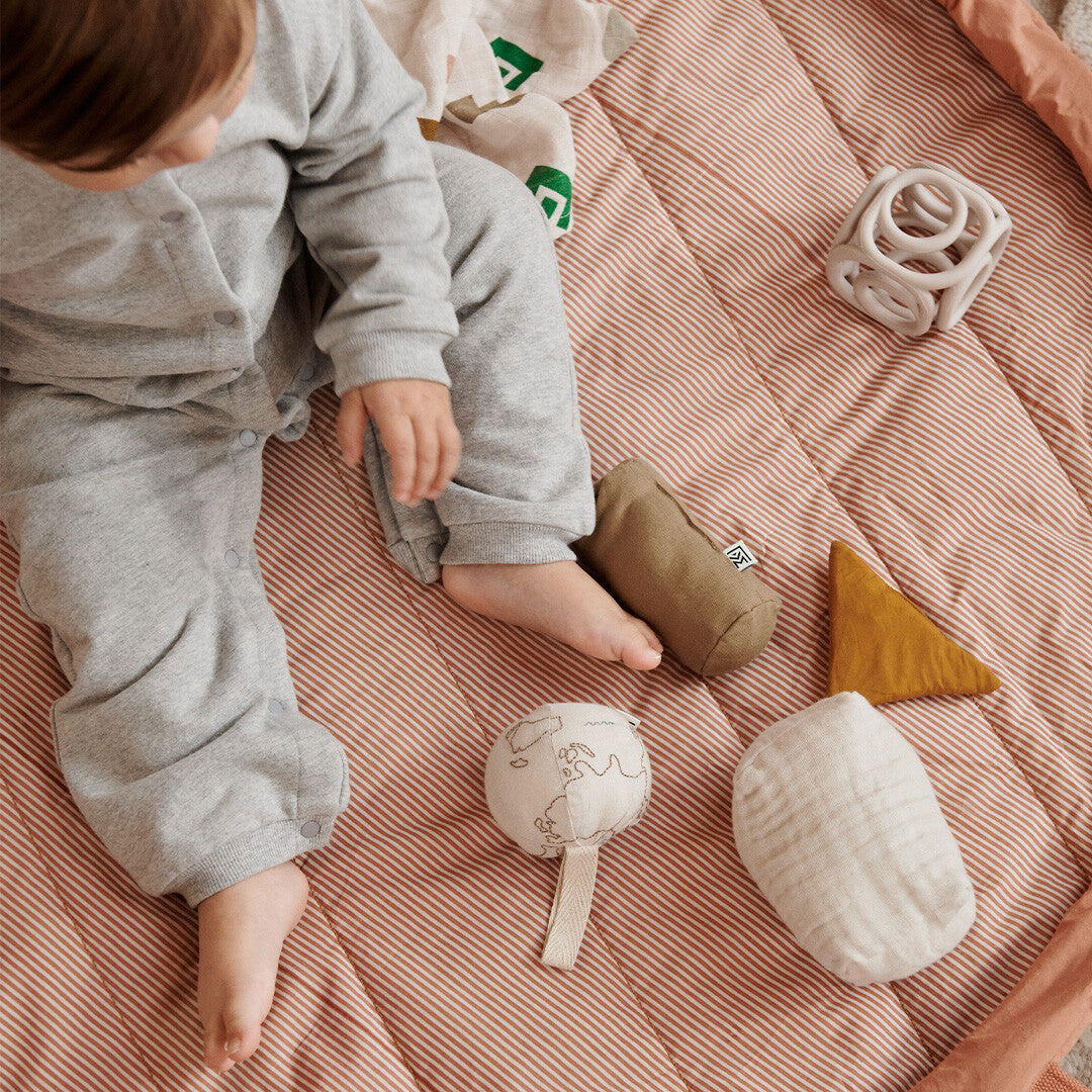 Een baby speelt op een gestreepte mat met verschillende speeltjes, waaronder het Liewood beige siliconen bijtspeelgoed.