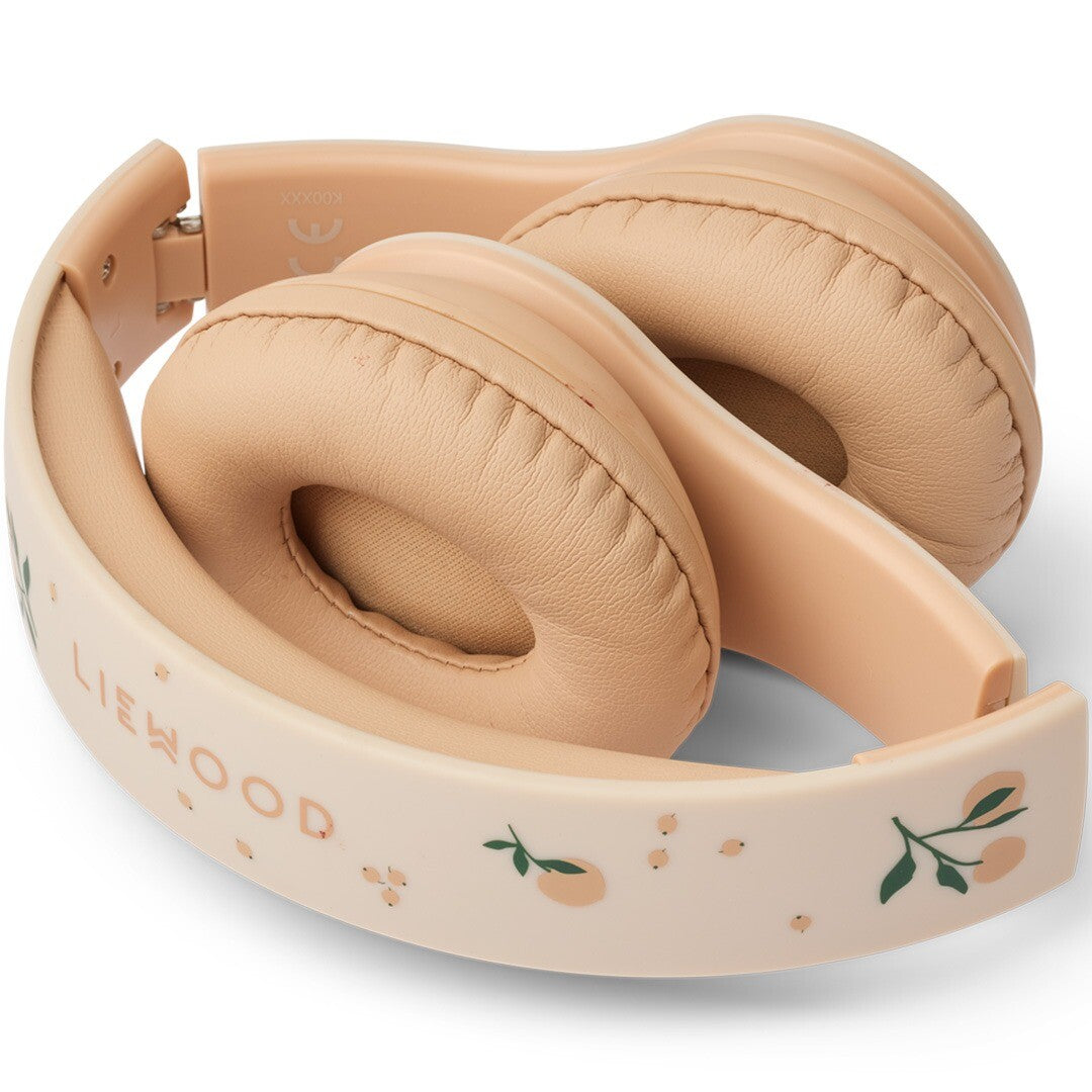 De opgevouwen Liewood draadloze kinderkoptelefoon in beige met perzikjespatroon, met het merklogo op de hoofdband.