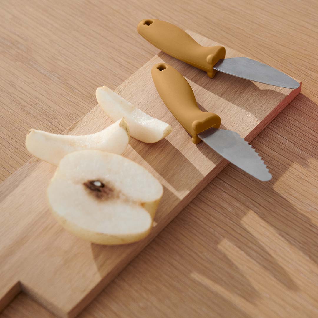 Twee Liewood kindermessen met okergele siliconen handvatten liggen naast gesneden peer op een houten snijplank.