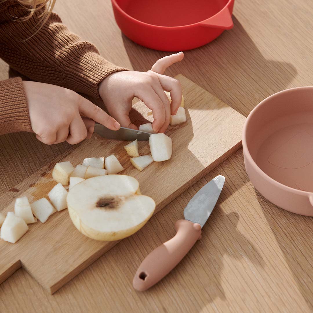 Een kind snijdt met een Liewood kindermes met roze handvat stukjes peer op een houten snijplank.