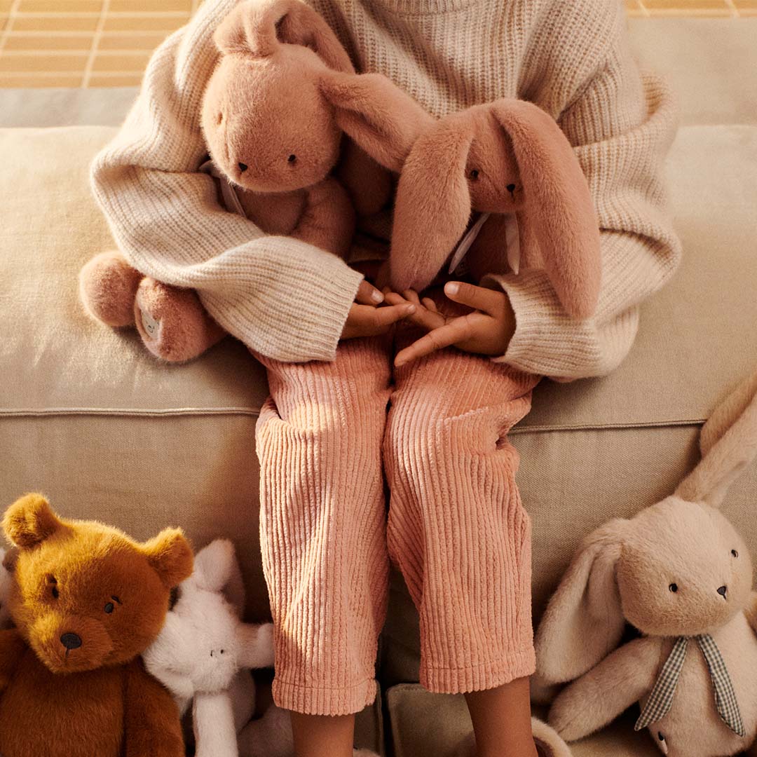 Een kind houdt twee roze pluchen knuffelkonijnen vast, omringd door andere knuffels.