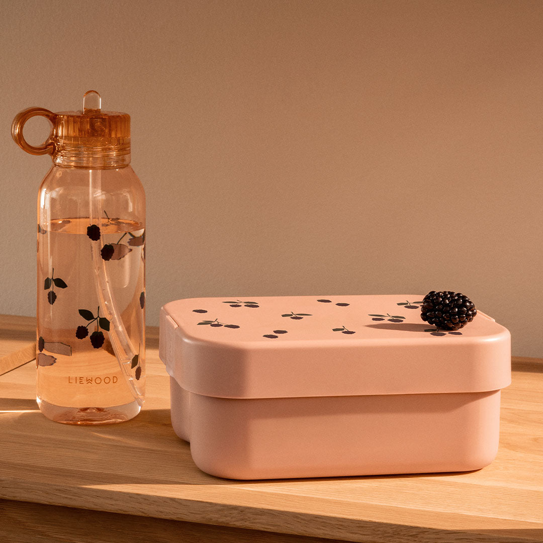 Liewood lunchset voor kinderen met roze broodtrommel en drinkfles met bessenpatroon, staand op een houten ondergrond met een braam op de trommel.