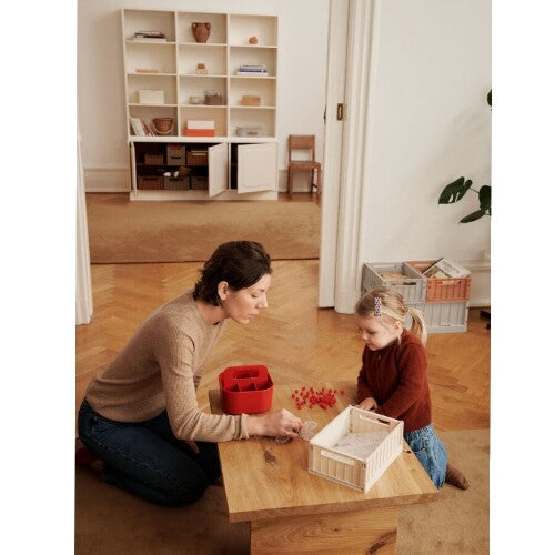 Een vrouw en een kind spelen met kralen, met een beige Liewood opbergkrat op een houten tafel.