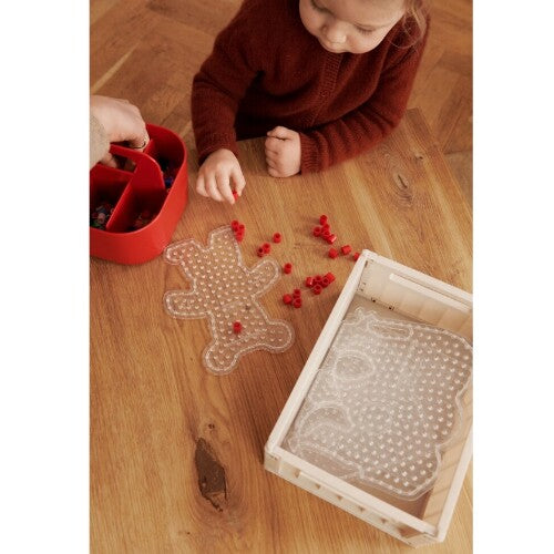 Detail van handen die met rode kralen en een beading board spelen, met een beige Liewood opbergkrat.