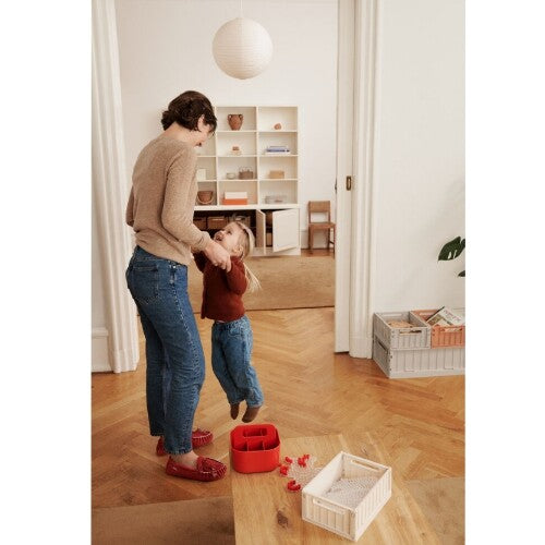 Een vrouw tilt een kind op in een woonkamer, met een beige Liewood opbergkrat op een houten tafel.