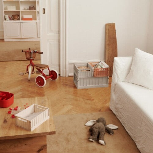 Een woonkamer met een rode driewieler en een beige Liewood opbergkrat op een houten tafel.