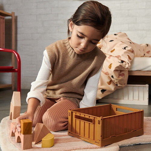 Een kind speelt met houten blokken naast een open bruine Liewood opbergkrat.