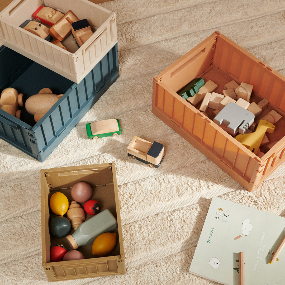 Drie Liewood opbergkratten in verschillende kleuren gevuld met houten speelgoed op een lichte vloer.