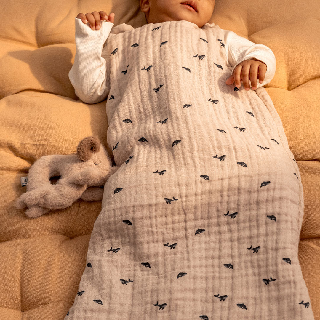 Een baby ligt in een slaapzak met de Liewood beige pluche olifant rammelaar ernaast.
