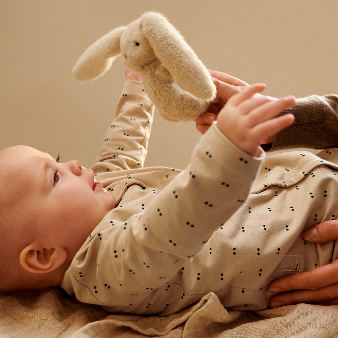 Een baby kijkt omhoog naar de Liewood beige pluche konijn rammelaar die door volwassen handen wordt vastgehouden.