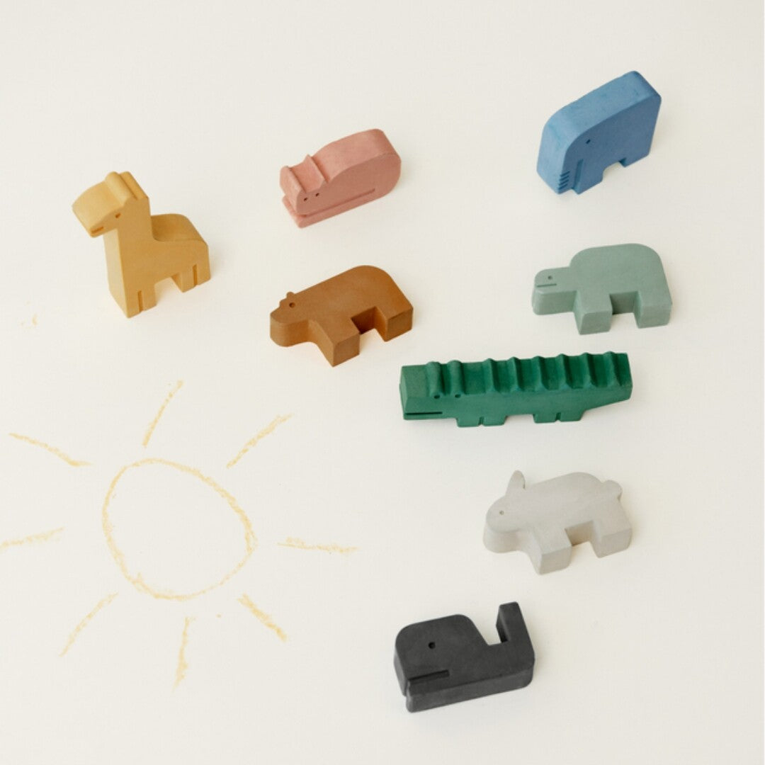 Acht gekleurde schoolkrijtjes in de vorm van dieren liggen verspreid op een lichte ondergrond met een getekende zon.