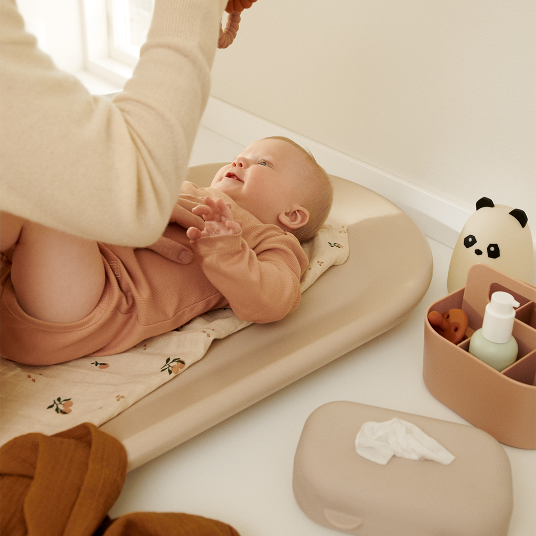 Een baby ligt op een verschoonkussen met de Liewood siliconen babydoekjes hoes en andere babyproducten ernaast.