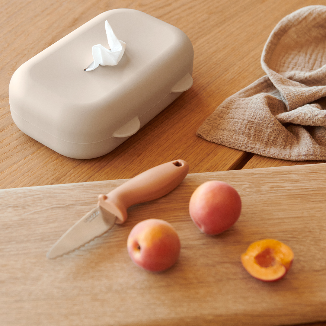 De Liewood siliconen babydoekjes hoes in zandkleur met een doekje eruit op een houten tafel, naast fruit.