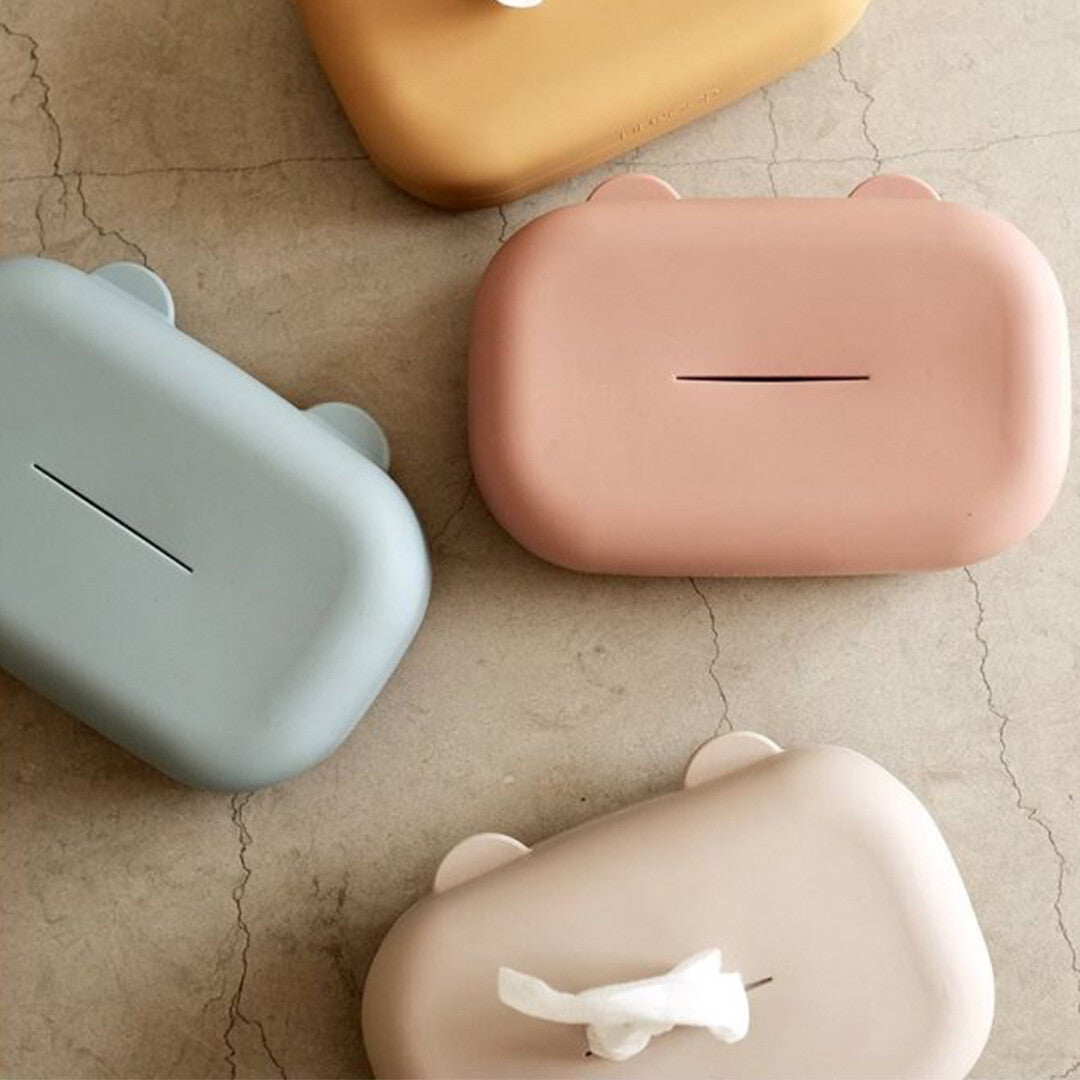 Vier Liewood siliconen babydoekjes hoezen in verschillende kleuren, waaronder zandkleur, roze en blauw, op een tegelvloer.