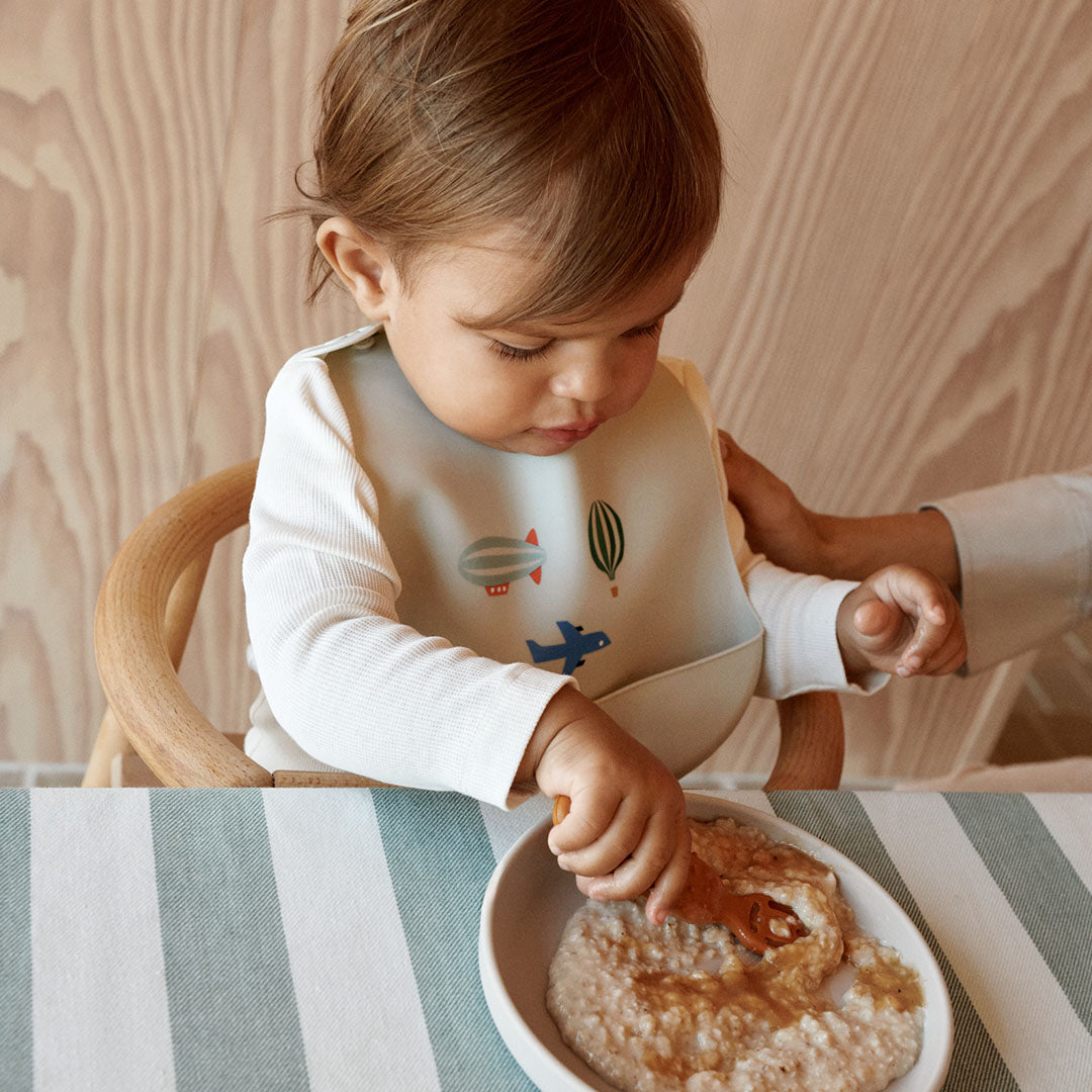 Een baby draagt een Liewood siliconen slabbetje met voertuigpatronen tijdens het eten.