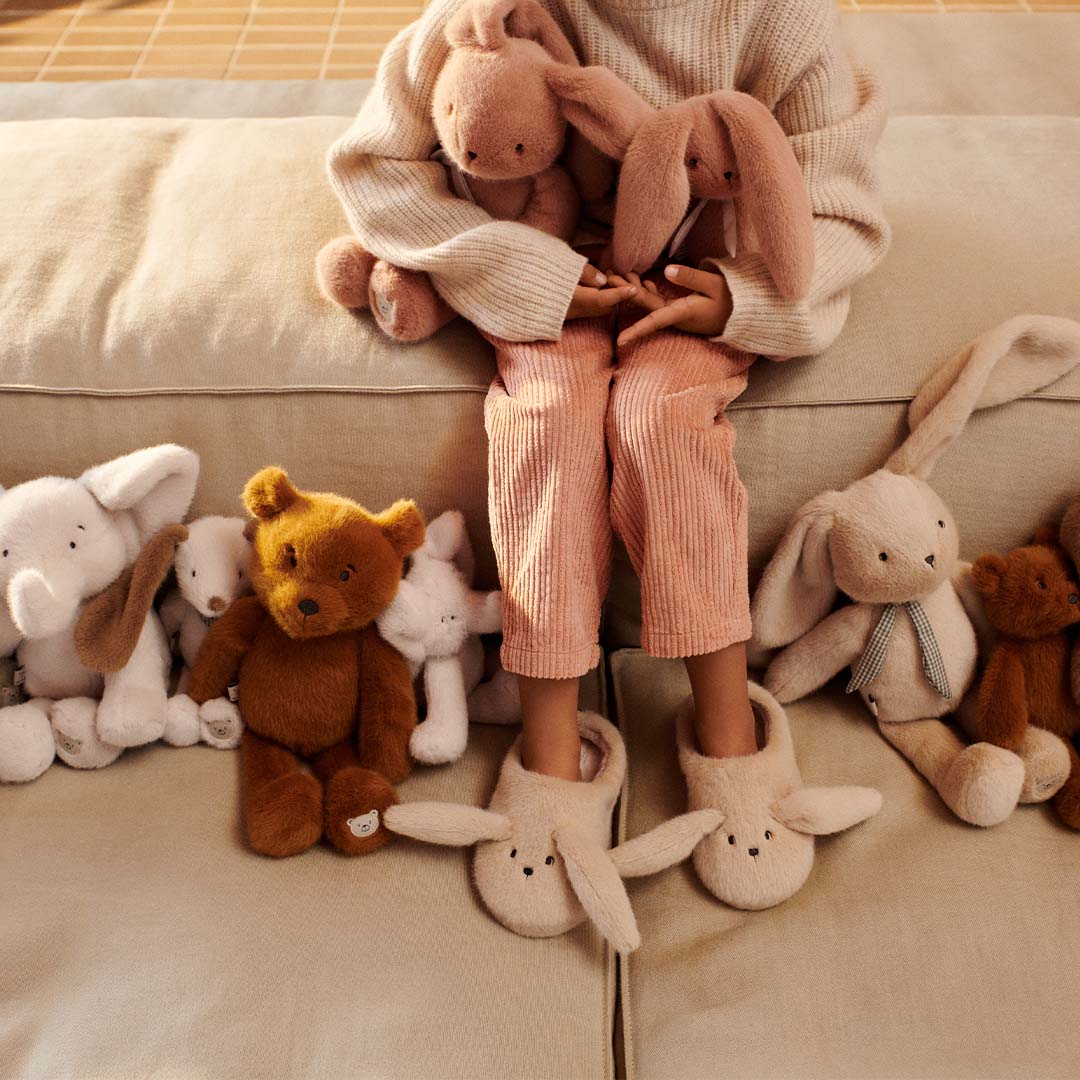 Een kind zit op een bank omringd door diverse pluchen dieren, waaronder de Liewood Berto teddybeer en konijnen.