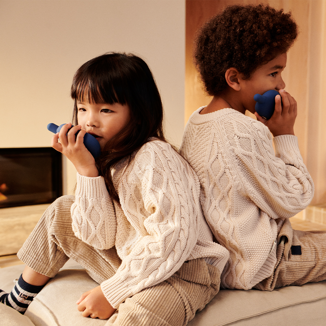 Twee kinderen spelen met Liewood indigo blauwe walkie talkies, zittend rug aan rug op een bank.