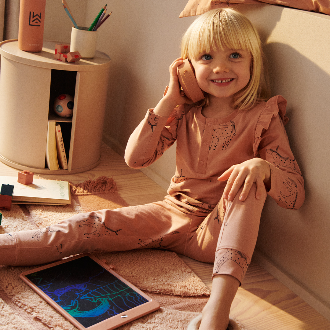 Een lachend meisje speelt met een beige Liewood walkie-talkie in een kinderkamer.