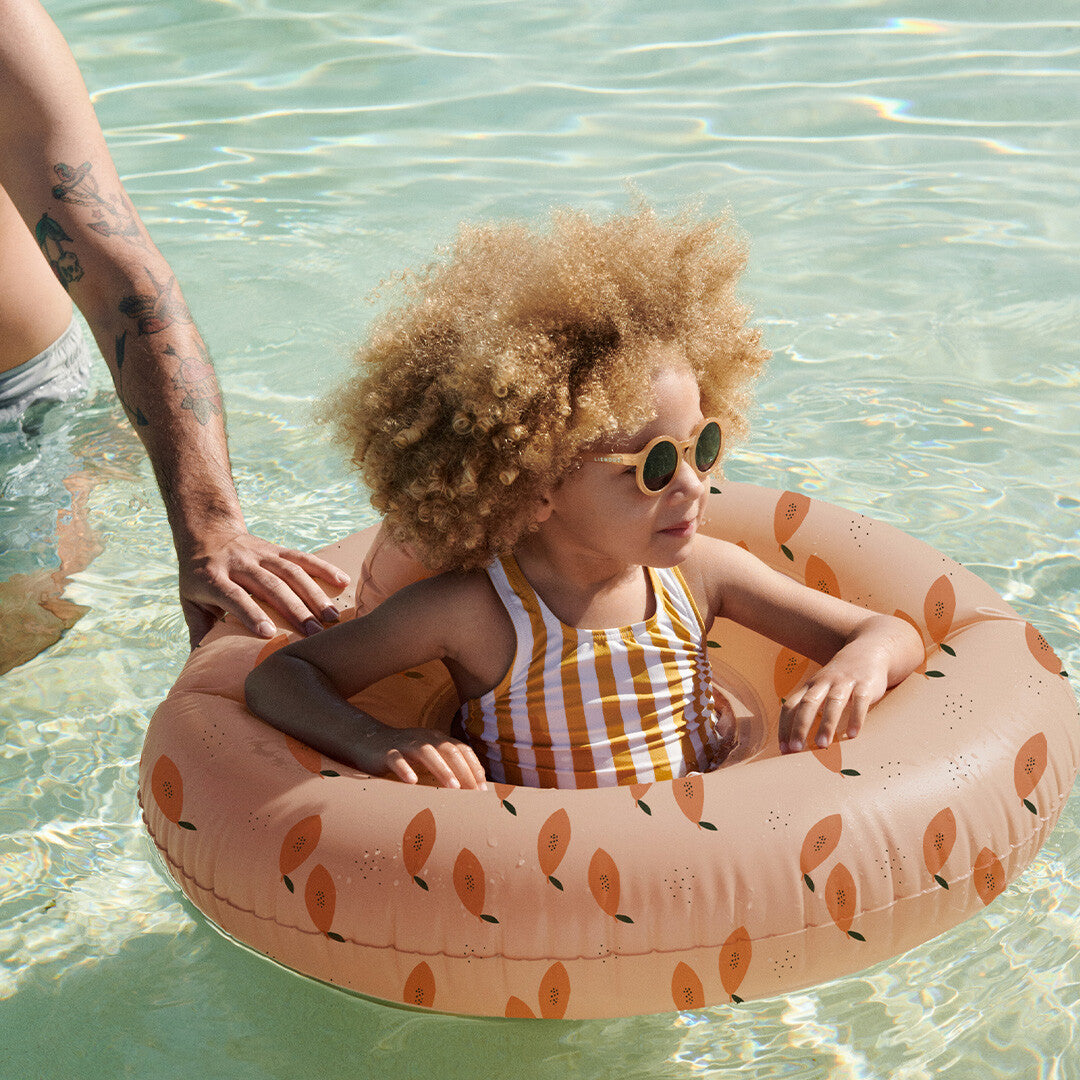 Een kind met zonnebril zit in een oranje zwemring in het water, vastgehouden door een volwassene.