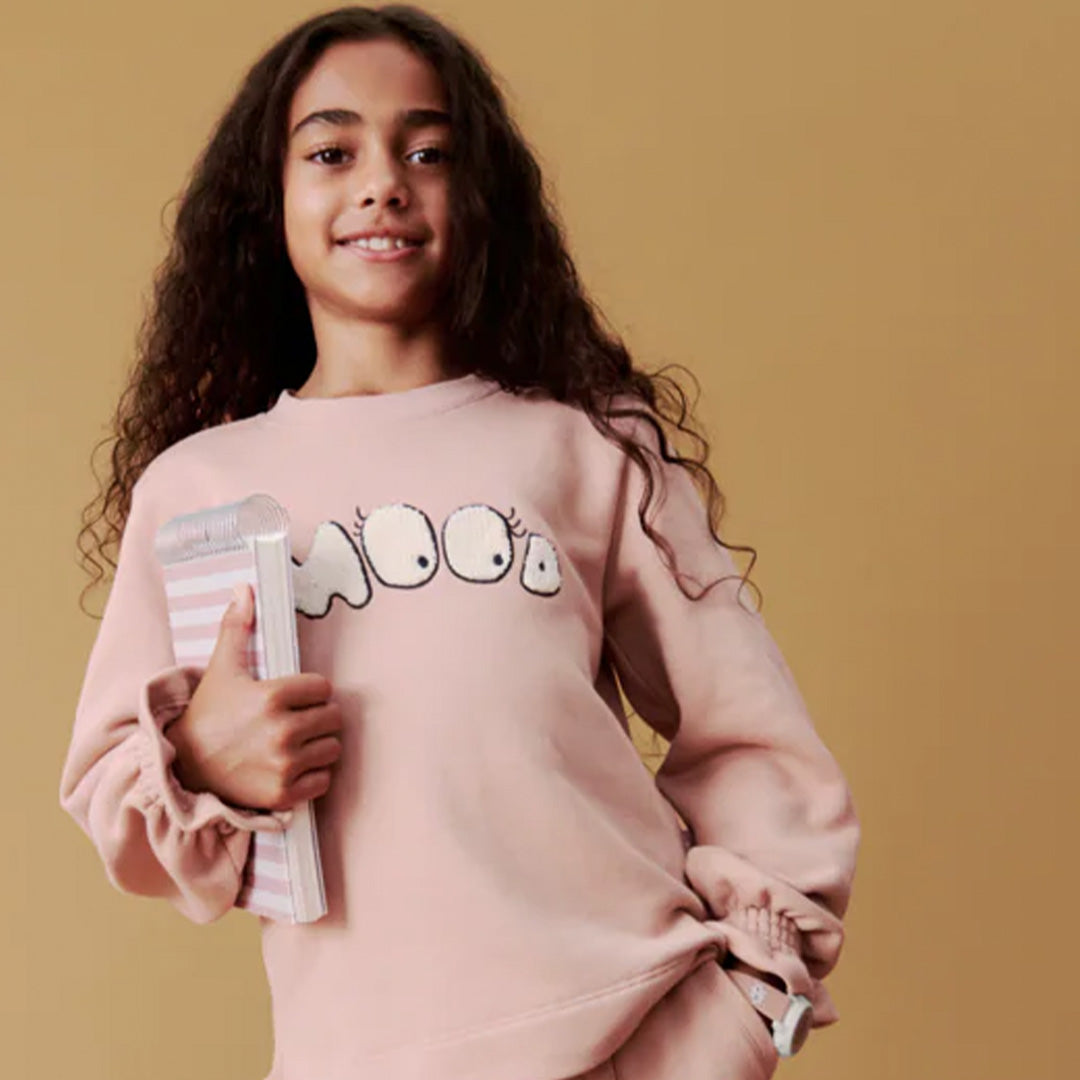 Een lachend meisje met lang krullend haar draagt een roze sweatshirt en het digitale kinderhorloge om haar pols.