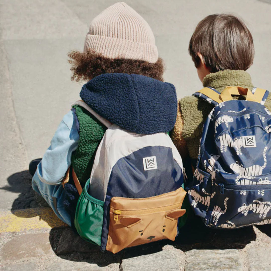 Twee kinderen zitten op de grond, waarvan één de Liewood peuterrugzak Andreas met berengezicht draagt.