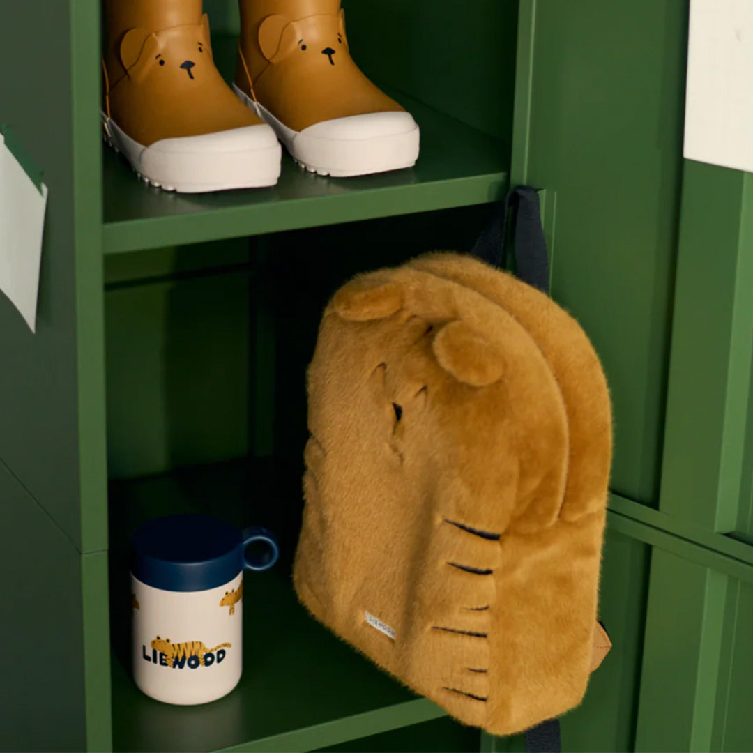 De bruine pluizige tijger peuterrugzak hangt in een groene locker, naast een beker en onder laarzen.