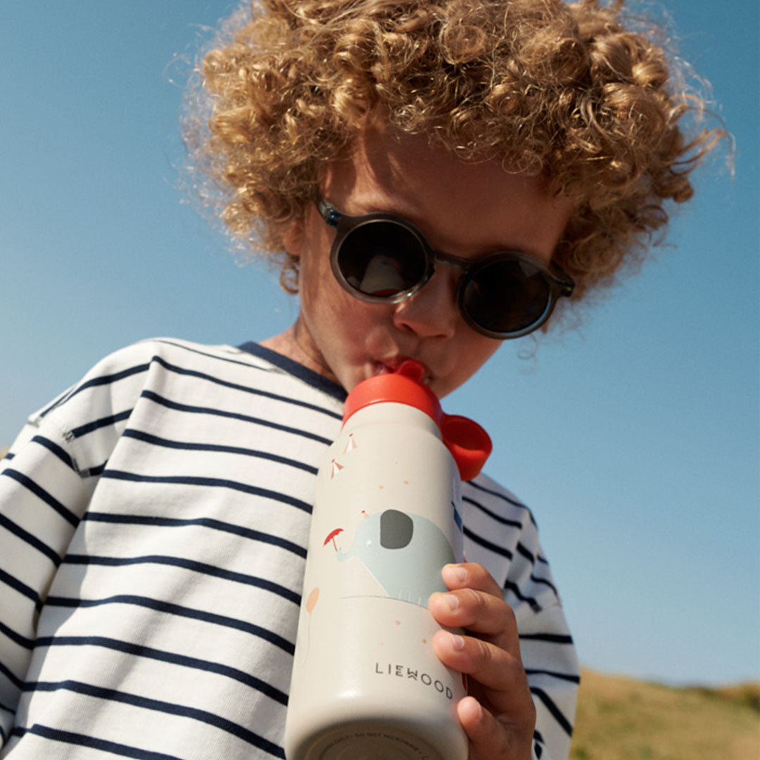 Een kind met zonnebril drinkt uit een Liewood kinder waterfles met olifantpatroon.