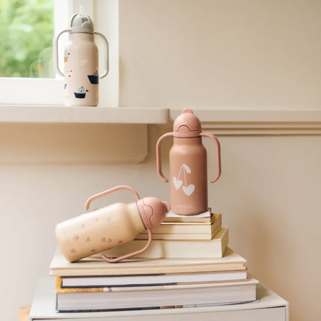 Drie Liewood baby thermosflessen met handvatten, waarvan twee op een stapel boeken en één op een vensterbank.