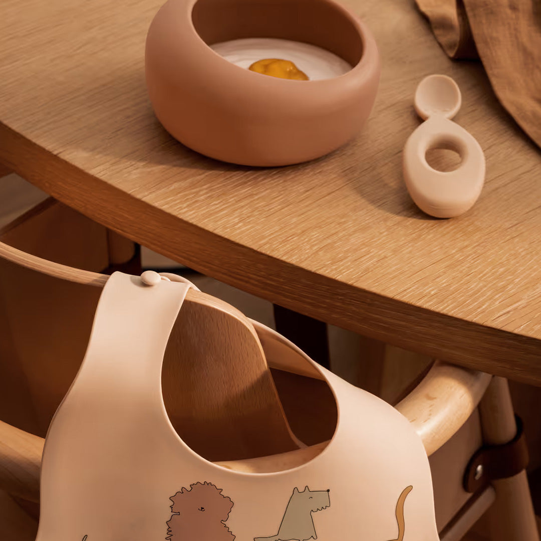 Liewood siliconen kinderkom met eten en lepel op een houten tafel, met een roze slabbetje op de voorgrond.
