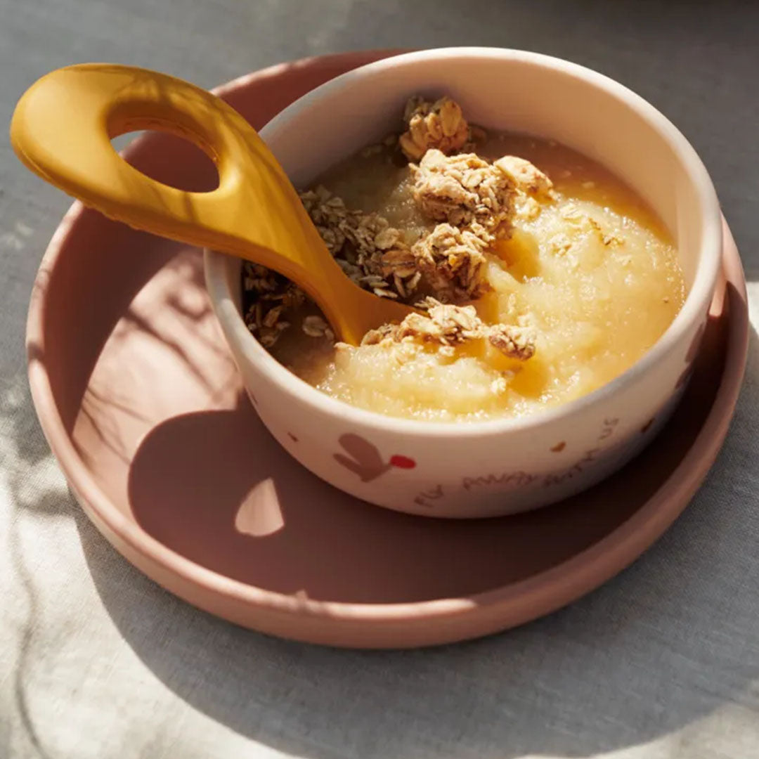 Roze siliconen kinderkom met vlinderpatroon gevuld met appelmoes en granola, met een gele lepel.