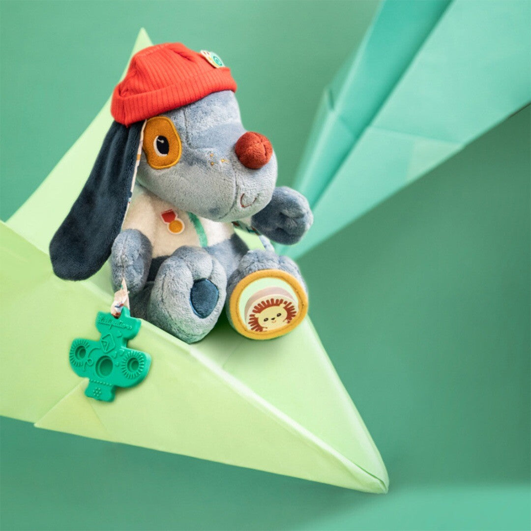 Lilliputiens activiteitenknuffel hond Jules zit op een papieren vliegtuig en houdt een groen vliegtuigje vast.