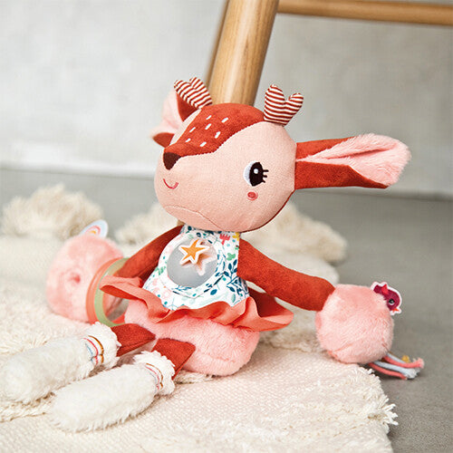 Lilliputiens activiteitenknuffel Stella, een bruin hertje met bloemenjurk en rammelaars, zittend op een wit kleed.