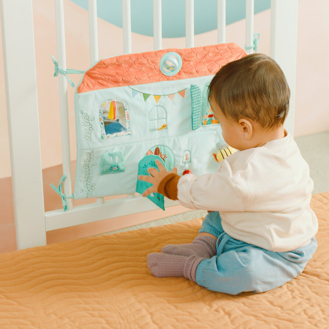 Een baby speelt met het Lilliputiens activiteitenpaneel dat aan een wit ledikant hangt.