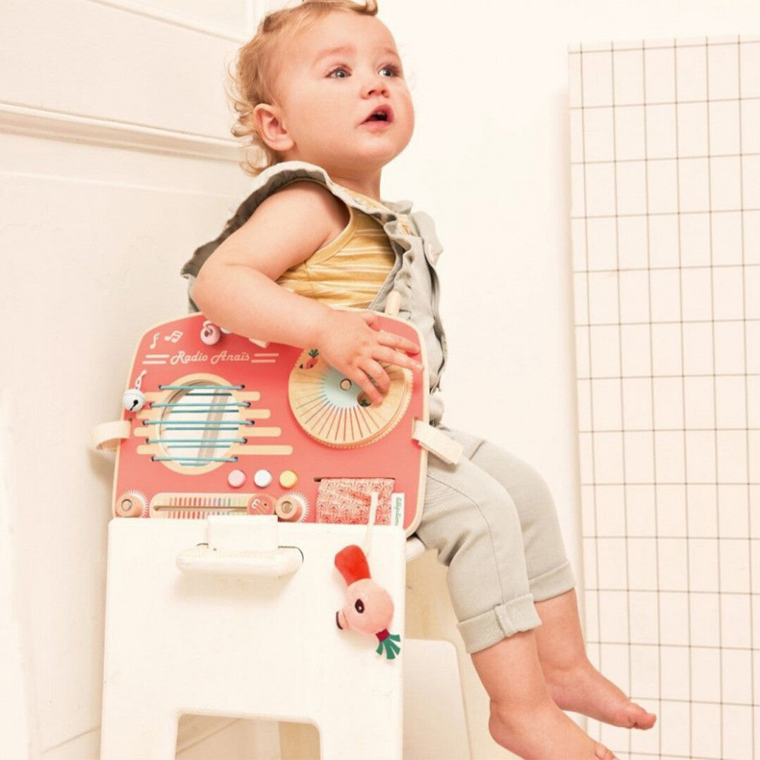 Een baby zit op een krukje en kijkt naar het Lilliputiens activiteitenpaneel in de vorm van een roze radio.