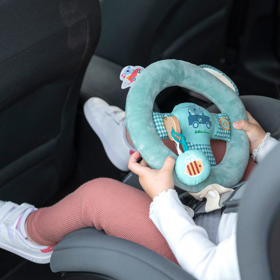 Een baby in een autostoel speelt met het Lilliputiens groene pluche activiteitenspeelgoed stuur.