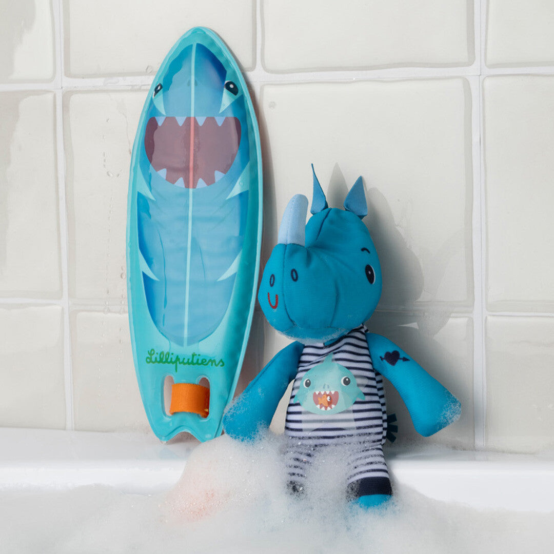 Lilliputiens badspeelgoed van een blauwe neushoorn knuffel en een haaien-surfplank in een bad met schuim.