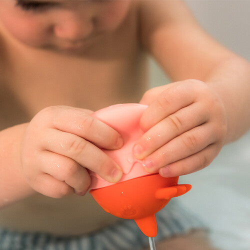 Kinderhanden knijpen in het Lilliputiens badspeeltje, waardoor water uit het oranje kuiken spuit.