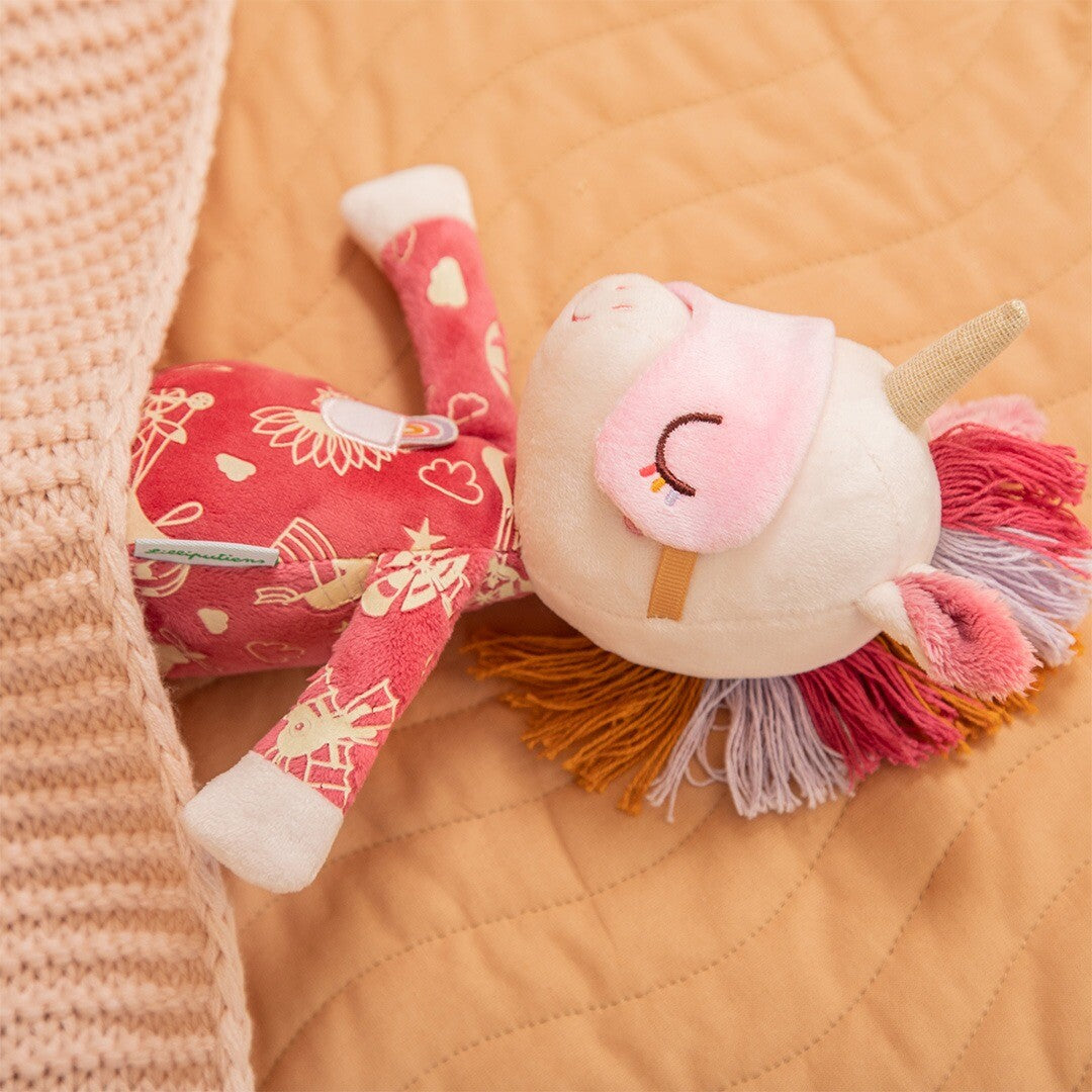 Pluchen eenhoorn knuffel Lena ligt op haar zij onder een lichtbruine deken.