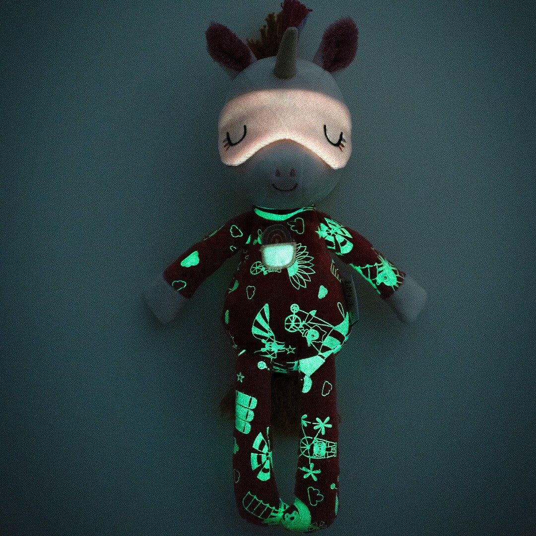 Pluchen eenhoorn knuffel Lena in het donker, waarbij de glow-in-the-dark patronen op haar pyjama en slaapmasker groen oplichten.