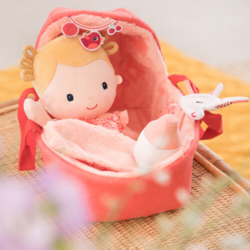 Een Lilliputiens lappenpop baby Lena in haar draagmand op een rieten krukje, met een flesje en eenhoorn knuffeltje ernaast.