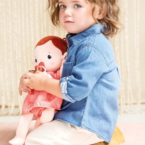 Een kind houdt de Lilliputiens lappenpop Rose met rode jurk en speen vast.